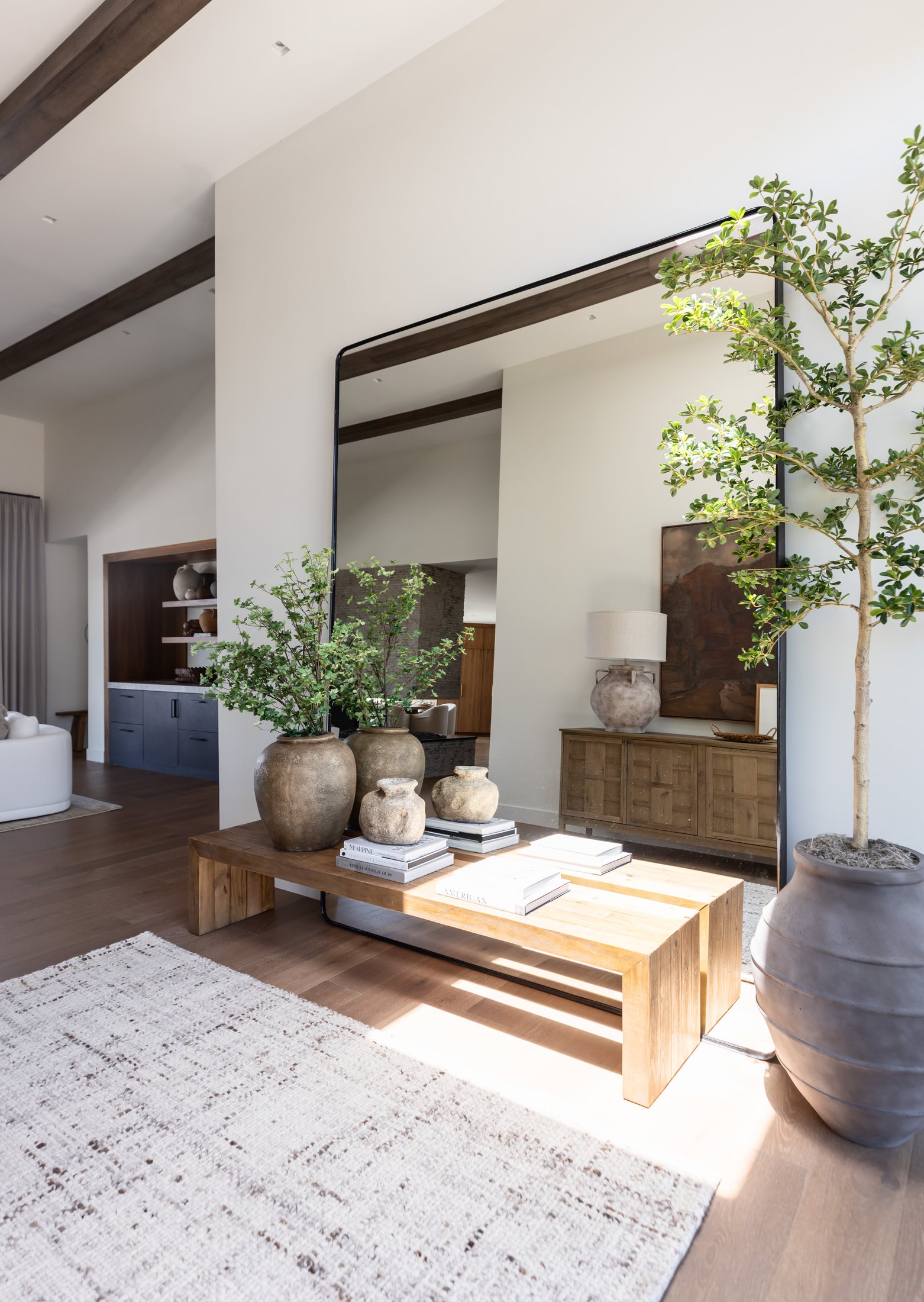 A sunlit organic-modern entry with a large black-framed mirror, olive tree, aged stone vessels and a wood bench by Kimberley Kay Interiors
