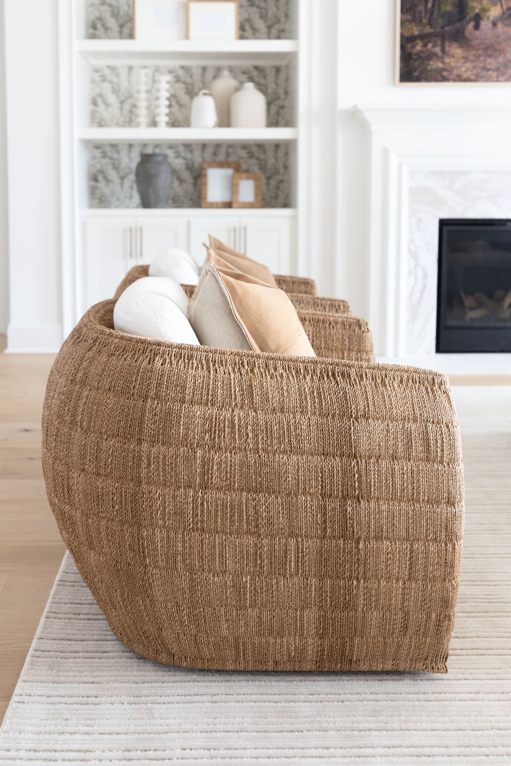 A woven barrel chair layered with linen pillows beside a marble fireplace and styled built-in shelves by Kimberley Kay Interiors